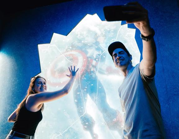 Two people are posing and taking a selfie with the Captain America trapped in ice statue at Marvel Earth's Mightiest Exhibition at Tākina in Wellington, New Zealand.