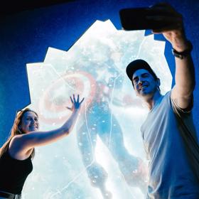 Two people are posing and taking a selfie with the Captain America trapped in ice statue at Marvel Earth's Mightiest Exhibition at Tākina in Wellington, New Zealand.