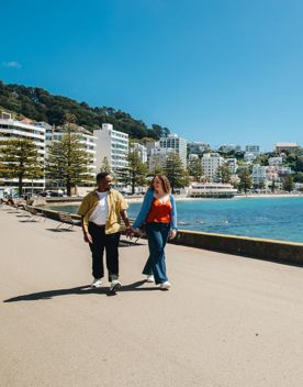 Two people stroll along Oriental Bay Parade in Wellington holding hands on a sunny day.