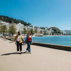 Two people stroll along Oriental Bay Parade in Wellington holding hands on a sunny day.