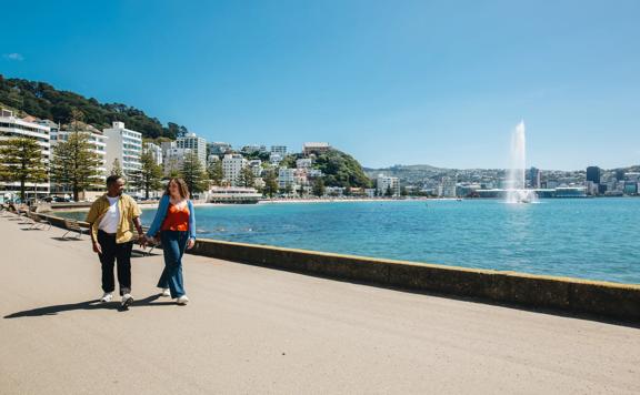 Two people stroll along Oriental Bay Parade in Wellington holding hands on a sunny day.