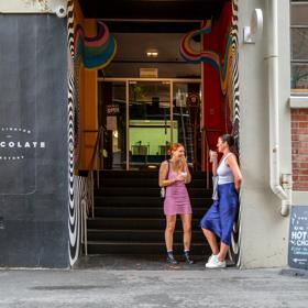 Outside the Wellington Chocolate Factory, 2 people stand enjoying a coffee. A large black wall is to the left with the business name on it.