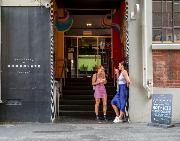 Outside the Wellington Chocolate Factory, 2 people stand enjoying a coffee. A large black wall is to the left with the business name on it.