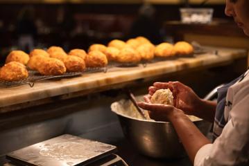 A baker at Pravda moulding a cheese scone, with a tray of baked scones under yellow light in the background.