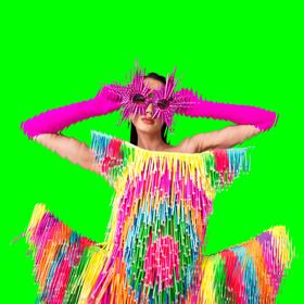 A model poses wearing a colourful fringe dress and hot pink sunglasses on a bright green background.