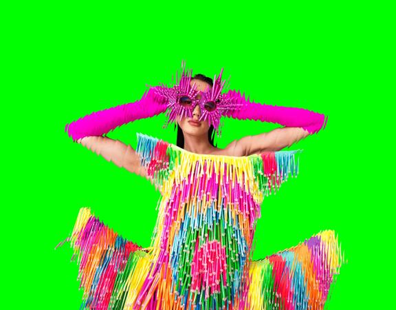 A model poses wearing a colourful fringe dress and hot pink sunglasses on a bright green background.