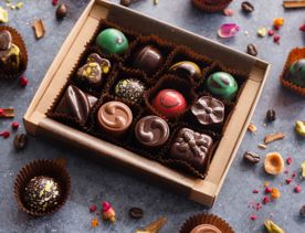 A box of artisanal chocolates from La Petite Chocolate, a chocolatier in Thorndon, Wellington. The box is on a grey-stone surface with colourful bits of ingredients and a few other chocolates arranged around it as decoration.
