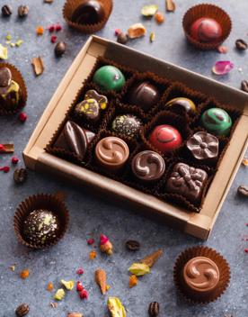 A box of artisanal chocolates from La Petite Chocolate, a chocolatier in Thorndon, Wellington. The box is on a grey-stone surface with colourful bits of ingredients and a few other chocolates arranged around it as decoration.