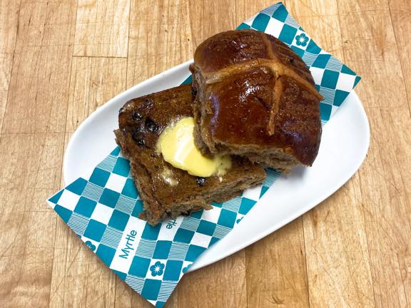 A single hot cross bun from Myrtle, a bakery in Mount Victoria, Wellington. The bun is cut into two pieces, top and bottom with a healthy slab of butter on the bottom piece. 