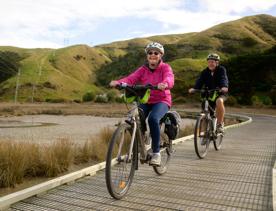 Two people riding bikes on Te Ara Piko Walkway. The trail features stretches of boardwalk and bridges, with amazing views of the harbour.