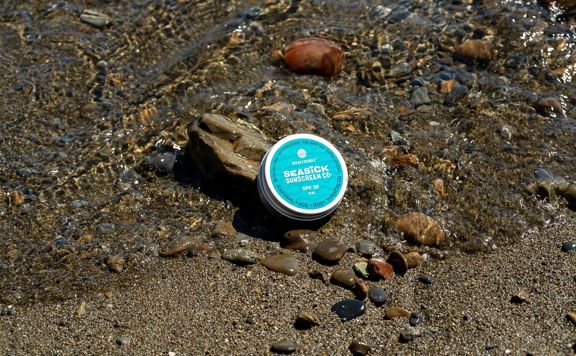 A tin of Seasick Sunscreen Co at the edge of the water on a pebbly beach.