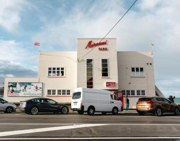 The exterior of Maranui Café, inside the Lyall Bay surf lifesaving club. The building is white with red accents and cars are parked outside.