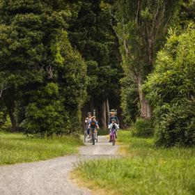 Two adults and two children riding bicycles on a gravel path surrounded by dense green trees and grass.