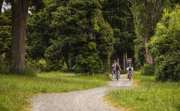 Two adults and two children riding bicycles on a gravel path surrounded by dense green trees and grass.