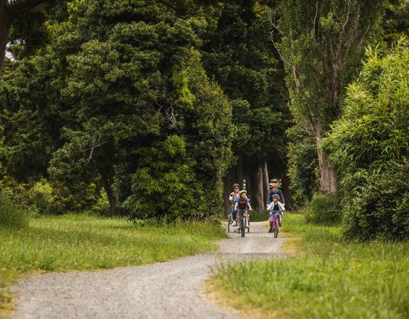 Two adults and two children riding bicycles on a gravel path surrounded by dense green trees and grass.