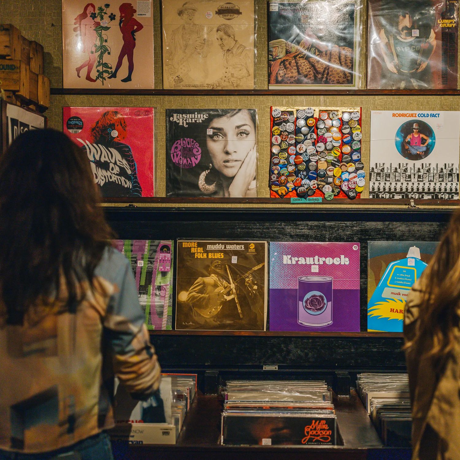 Two people stand facing a display of vinyl records in a record shop, browsing a wall filled with colourful album covers above bins of records.