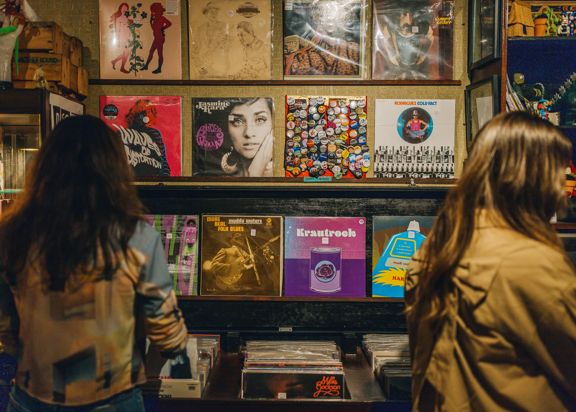 Two people stand facing a display of vinyl records in a record shop, browsing a wall filled with colourful album covers above bins of records.