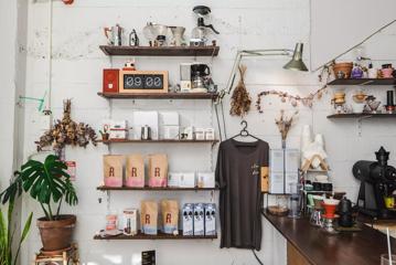 A white wall with wooden shelves inside a café. There are bags of coffee beans, oat milk, coffee apparatuses and other items displayed on the shelves.