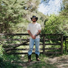 Kiwi musician, Te Wehi, stands in front of a wooden fence surrounded by trees.