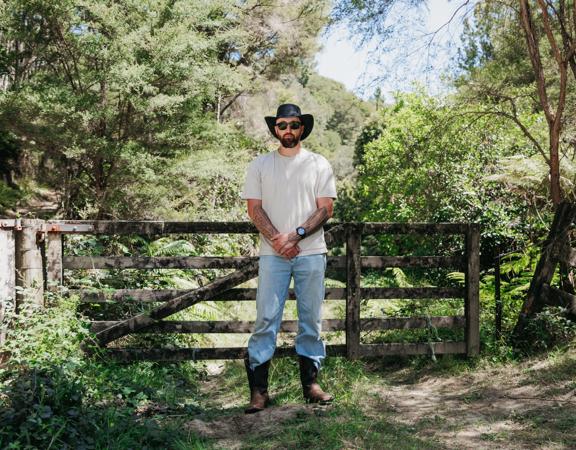 Kiwi musician, Te Wehi, stands in front of a wooden fence surrounded by trees.