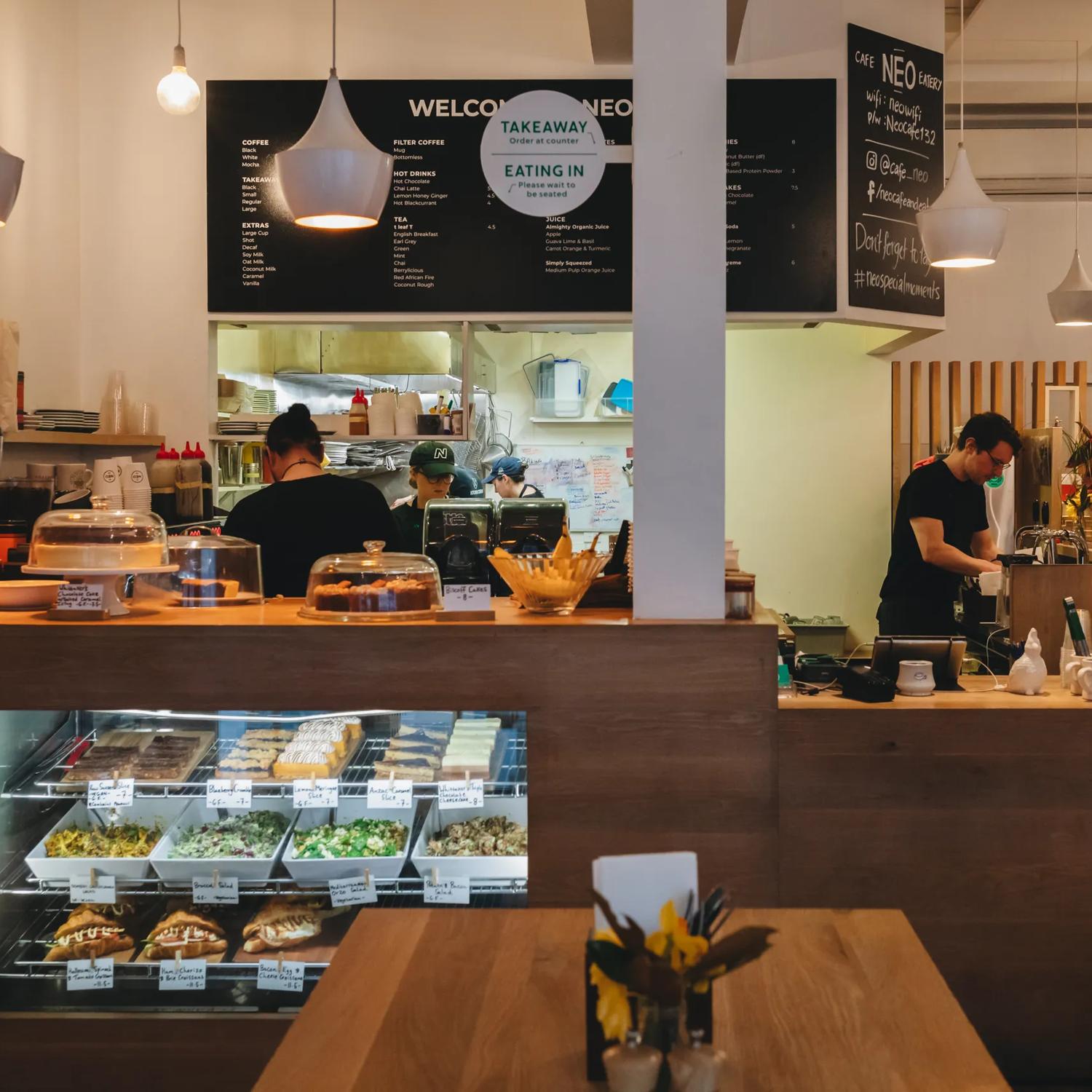 The front counter at Neo Café & Eatery in Te Aro, Wellington. Four people are working in the back and behind the counter, wooden furniture and pastries and salads for sale.