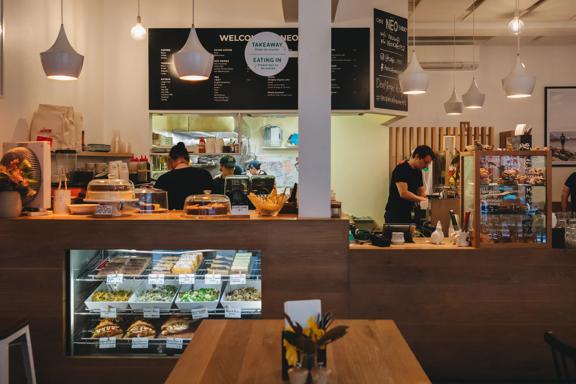 The front counter at Neo Café & Eatery in Te Aro, Wellington. Four people are working in the back and behind the counter, wooden furniture and pastries and salads for sale.