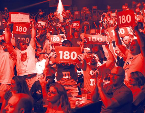 An excited audience cheering and holding red signs with the number "180" on them at the Professional Darts Corporation ANZ Premiere League competition.