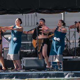 Five people wearing traditional Māori outfits, performing a dance on a stage.