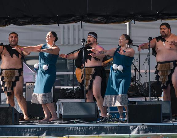 Five people wearing traditional Māori outfits, performing a dance on a stage.