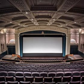 Inside a cinema inside Embassy Theatre. The large auditorium has high vaulted ceilings,  art deco reliefs on the walls and a large projection screen with open teal curtains. 