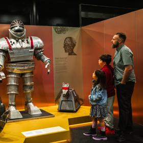 An adult and two children look at three robots on display at the Doctor Who Worlds of Wonder exhibition. 