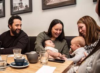 3 parents sat at a table inside Kāinga Eatery, holding 2 babies, and enjoying talking over their meals.