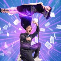 A magician crouching down with a woman floating over him and papers flying around them.
