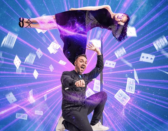 A magician crouching down with a woman floating over him and papers flying around them.