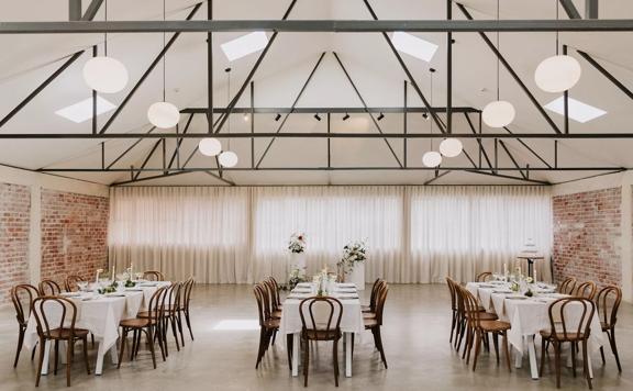The interior of Whisky and Wood, an event venue in Wellington Central, setup with three banquet tables, with white table clothes and eight wooden chairs at each, exposed brick walls and circular lights suspended from the ceiling. 