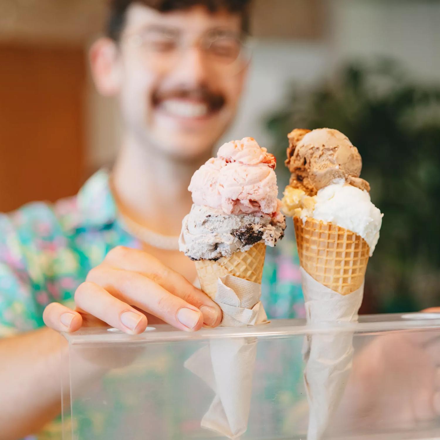 Two double scooped gelato in waffle ice cream cones set in a clear plastic stand at Raglan Roast Coffee at Chaffers Dock in Te Aro in Wellington. The smiling worker is our of focus but visible behind the ice cream. 