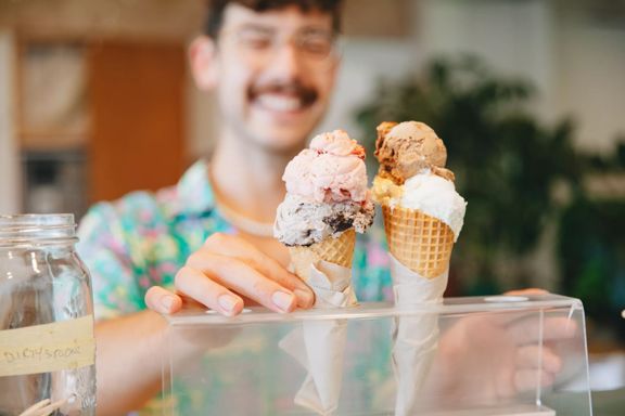 Two double scooped gelato in waffle ice cream cones set in a clear plastic stand at Raglan Roast Coffee at Chaffers Dock in Te Aro in Wellington. The smiling worker is our of focus but visible behind the ice cream.