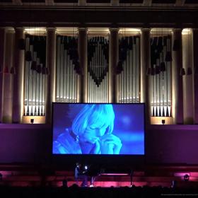 A projector screen displaying a scene from the video game Final Fantasy is set up on stage in a dark auditorium with a pianist playing an accompaniment.