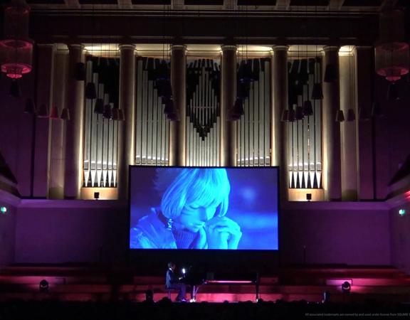 A projector screen displaying a scene from the video game Final Fantasy is set up on stage in a dark auditorium with a pianist playing an accompaniment.