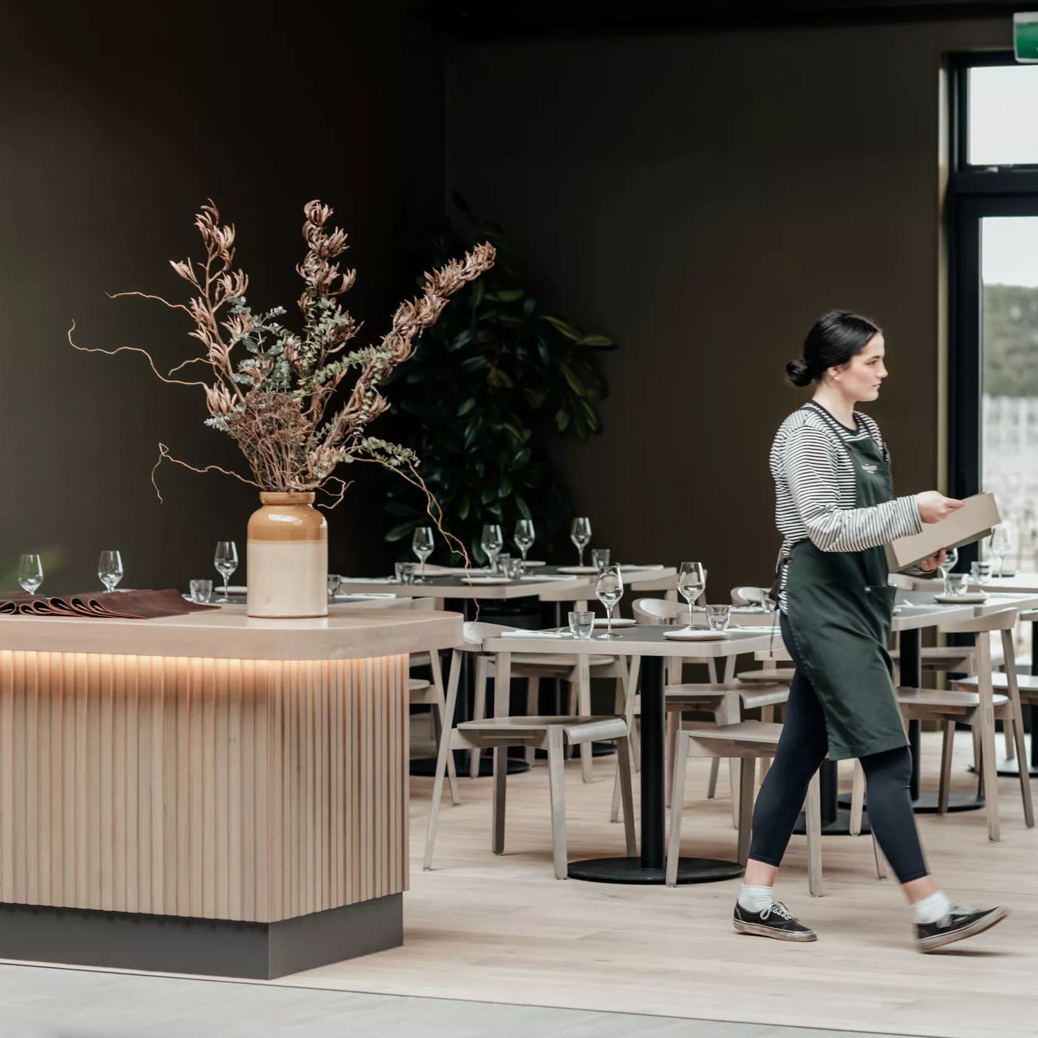 A server wearing a striped shirt and green apron walks across the empty, set dining room at The Runholder, an upscale restaurant in Martinborough.