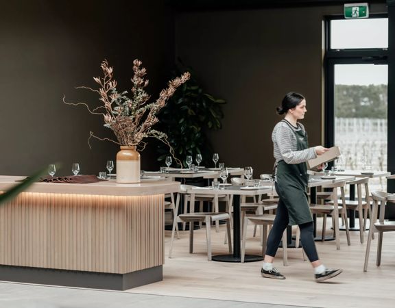 A server wearing a striped shirt and green apron walks across the empty, set dining room at The Runholder, an upscale restaurant in Martinborough.