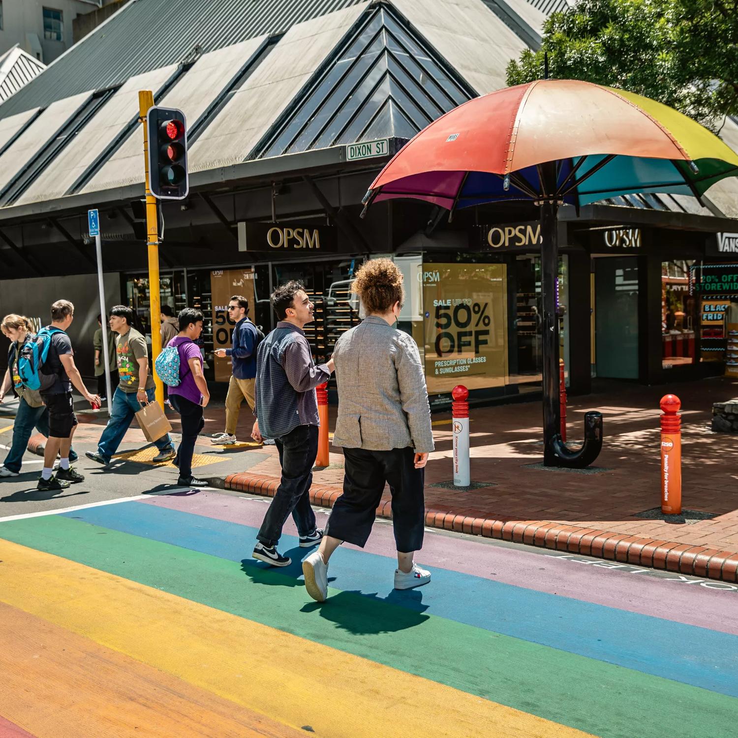 2 people walking across the rainbow crossing on Cuba street, next to the rainbow umbrella.