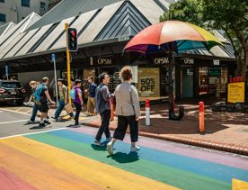 2 people walking across the rainbow crossing on Cuba street, next to the rainbow umbrella.