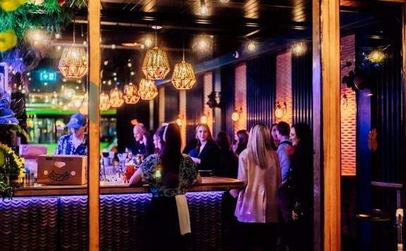 Looking into the open glass doors at Lulu Bar, workers and patrons are standing by the counter in the dark, amber-lit space.