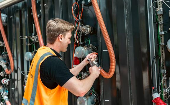 A person wearing an orange safety vest, using a tool to work on a piece of equipment as a part of the Summer of Engineering internship programme.