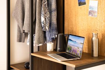 A close-up of the desk and wardrobe arrangement in a Haka House private room. There is a laptop, kettle and drinks on the desk, with clothes hanging in the wooden wardrobe.