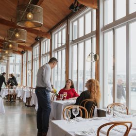 Two people enjoying a meal at a fine dining restaurant with white tablecloths. A waiter is showing them a bottle of wine.