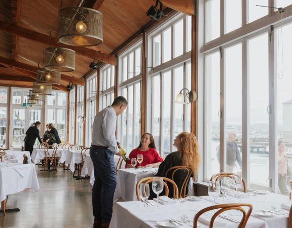 Two people enjoying a meal at a fine dining restaurant with white tablecloths. A waiter is showing them a bottle of wine.
