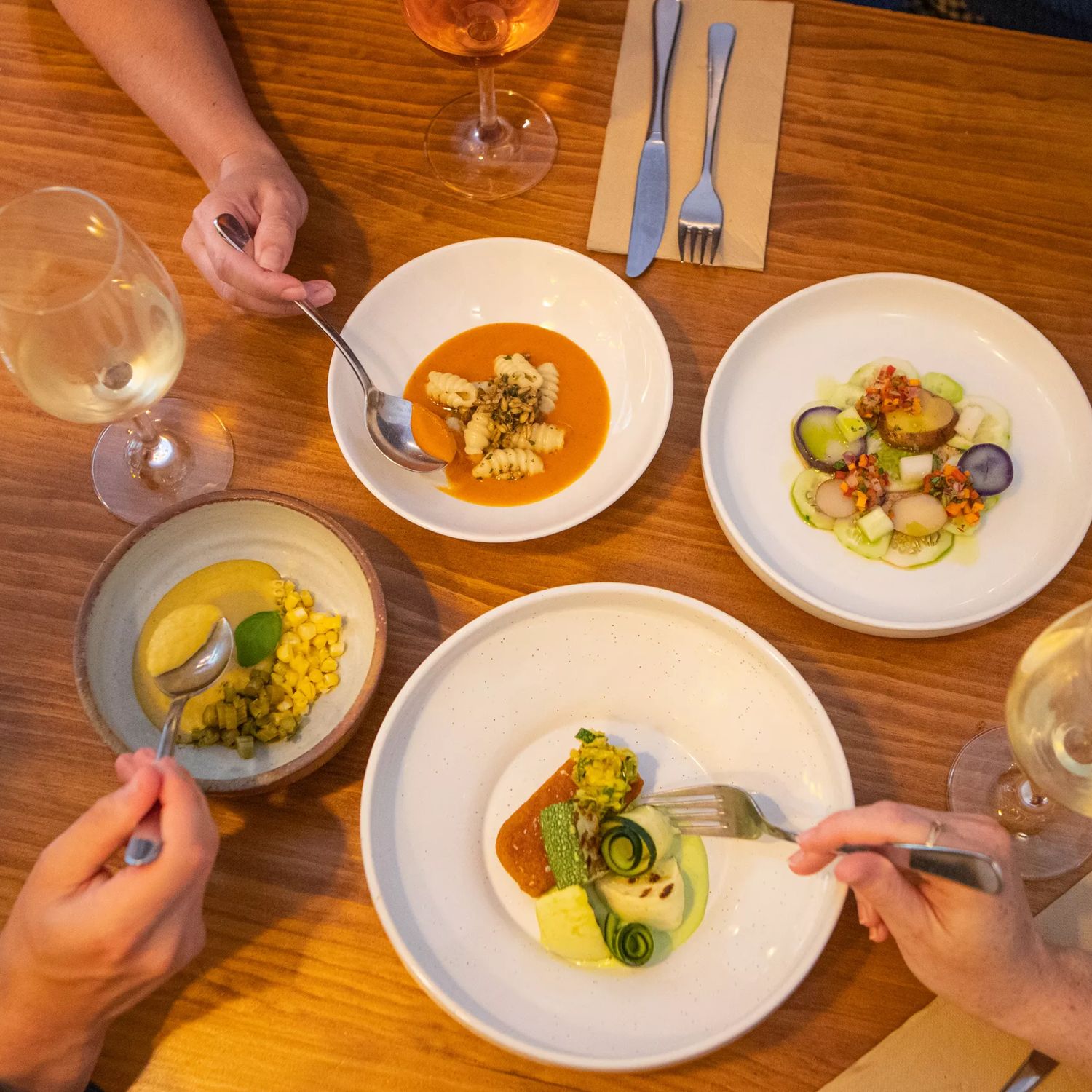 Four fine dining dishes on a table with people's hands reaching in with utensils.