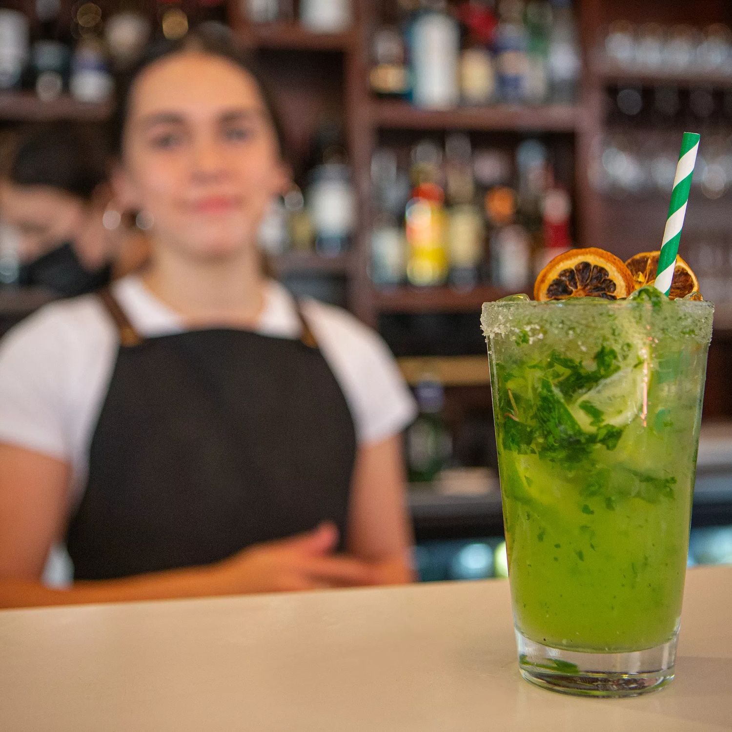 A green cocktail sits towards the camera with a waiter blurred out in the background, and Long beach.